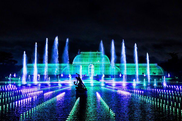 فواره های بلند و نورپردازی خطی رنگی روی آب در برابر گلخانه شیشه ای روشن شده به رنگ سبز در کیو گاردنز