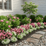 باغچه کوچک در حیاط خانه با لایه بندی گل‌های صورتی و سفید، درختچه‌های سبز در پس‌زمینه و مسیر سنگی با قلوه سنگ‌های طبیعی کنار دیوار خانه