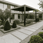 نمای کلی محوطه سازی ویلا با پرگولای فلزی، مسیرهای اسلب روشن، قاب بندی باغچه ها با سنگریزه و چیدمان مینیمال گیاهان