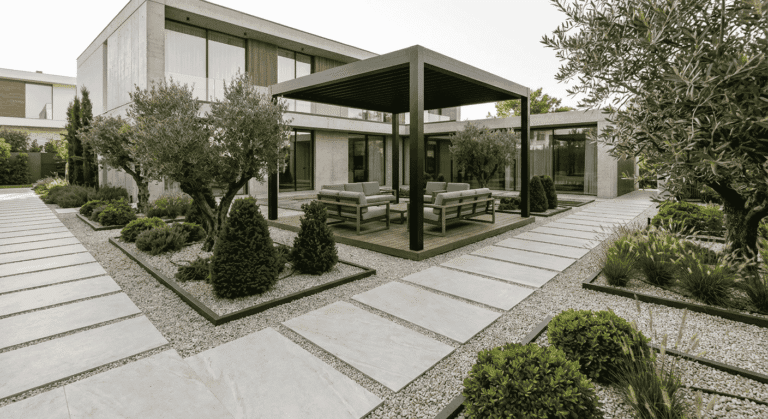 نمای کلی محوطه سازی ویلا با پرگولای فلزی، مسیرهای اسلب روشن، قاب بندی باغچه ها با سنگریزه و چیدمان مینیمال گیاهان