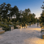 مسیر سنگفرش پارک در غروب با نیمکت های چوبی، چراغ های کوتاه بولارد و تردد عابران و دوچرخه سوارها