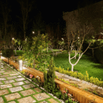 نورپردازی مسیر سنگ‌فرش باغ ویلا با استفاده از چراغ‌های ایستاده و تزئینات چوبی برای زیبایی و امنیت در شب